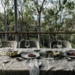 A beautifully set outdoor dining table with gourmet dishes and wine, surrounded by nature.