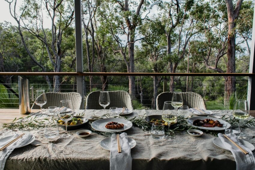 A beautifully set outdoor dining table with gourmet dishes and wine, surrounded by nature.